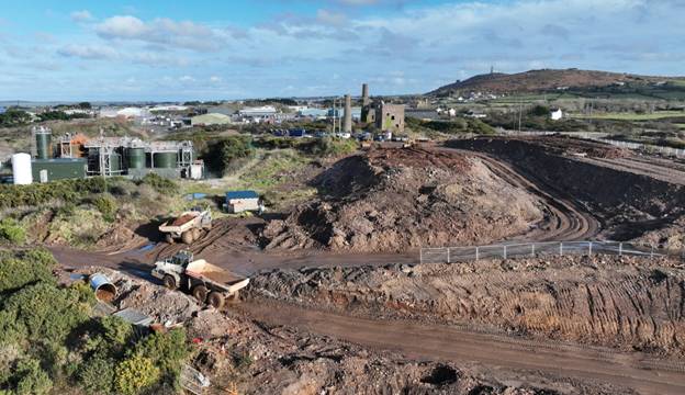 The image shows a construction site with heavy machinery and excavation equipment, including a large dump truck and a cement mixer, surrounded by barriers and under a partly cloudy sky. AI-generated content may be incorrect.