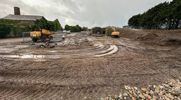 The image shows a construction site with a yellow excavator, dirt, and some buildings under a cloudy sky. AI-generated content may be incorrect.