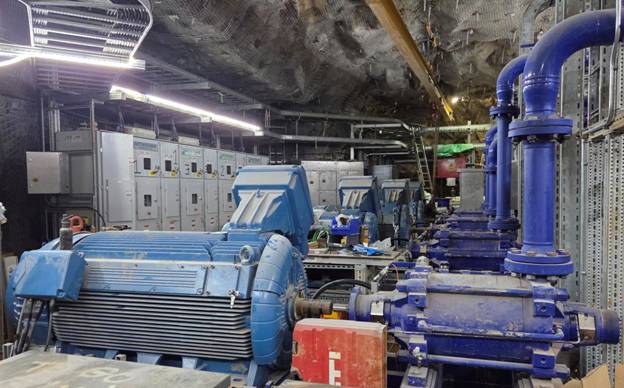 The image depicts an industrial setting with a series of large, blue hydraulic machinery and equipment, possibly for manufacturing or processing, situated in a dimly lit, underground environment. AI-generated content may be incorrect.