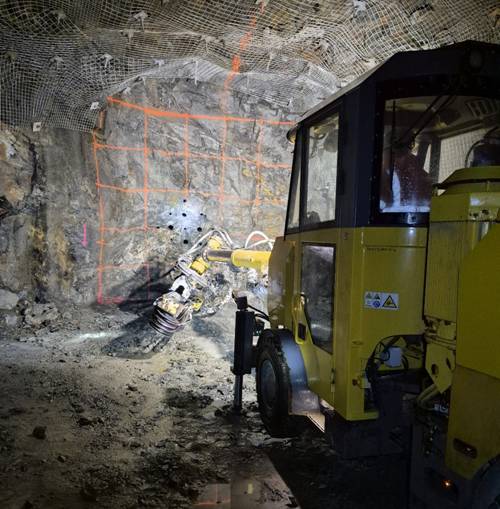The image depicts a construction site with a yellow bulldozer at the entrance of a tunnel, surrounded by marked measurements and a wall with visible construction activity. AI-generated content may be incorrect.