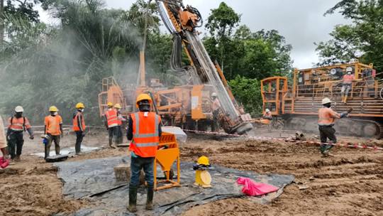 A group of people in orange vests and helmets standing in a dirt area Description automatically generated