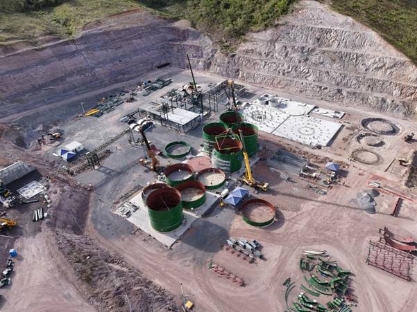 The image depicts an industrial construction site with several large, green cylindrical tanks and heavy machinery, surrounded by a dirt and rocky terrain. AI-generated content may be incorrect.