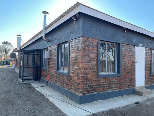 The image shows a small, modern brick house with blackout windows, a black metal gate, and a black roof, situated on a grassy, barren plot under a clear blue sky. AI-generated content may be incorrect.