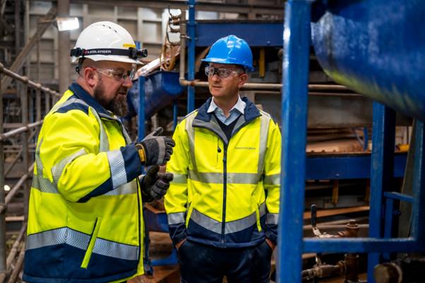 Tungsten West begins production at Hemerdon mine (TUN) The image shows two workers wearing high-visibility safety vests and hard hats, engaged in some form of construction or maintenance work, possibly operating or inspecting large industrial machinery or equipment. AI-generated content may be incorrect.