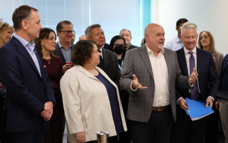 The image depicts a group of people standing in a professional setting, possibly a meeting or a conference, with some holding documents or presentations, and a few individuals engaged in conversation.

AI-generated content may be incorrect.