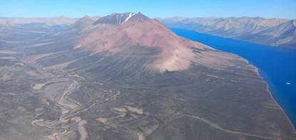 The image depicts a vast, barren, volcanic landscape with a prominent, red-colored mountain and a clear, expansive body of water in the background. AI-generated content may be incorrect.