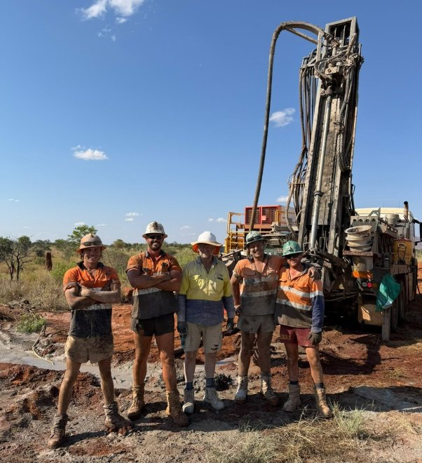 The image shows a group of five people standing together on a dirt road, wearing construction hard hats and vests, with a large construction vehicle in the background.
AI-generated content may be incorrect.
